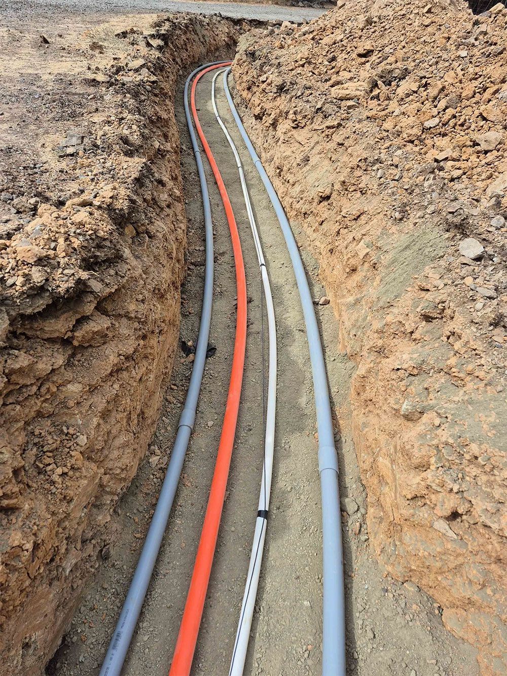 Trenched cables: Orange, white, and gray conduits are laid in a brown earthen ditch, ready for burial.