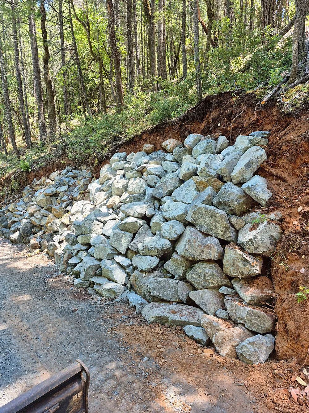 Rock retaining wall on a hillside next to a gravel road in a wooded area.