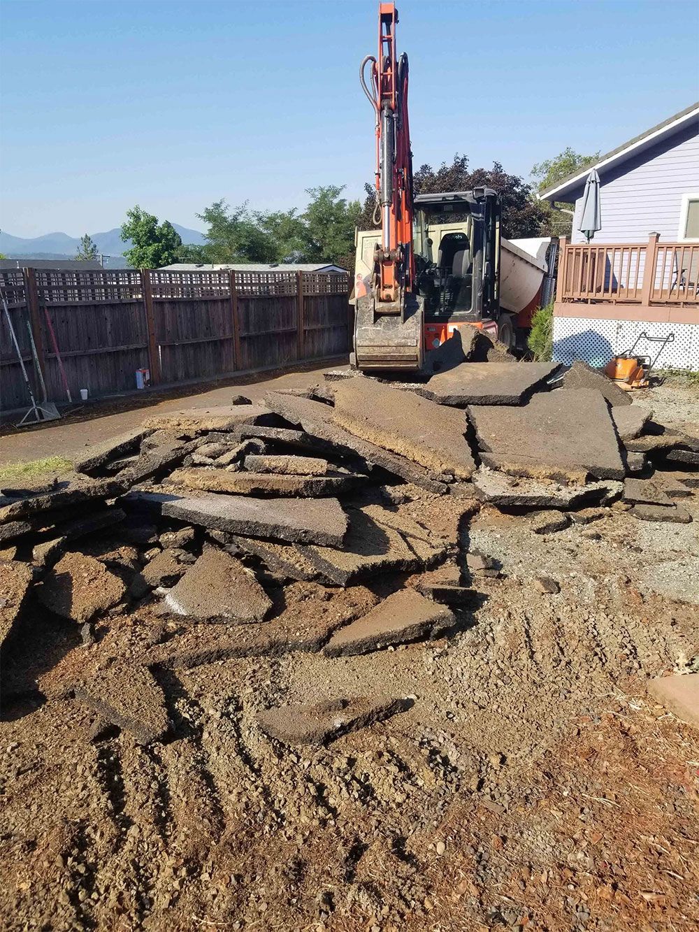 Mini excavator removing asphalt slabs in a residential yard. Brown wood fence and house in background.