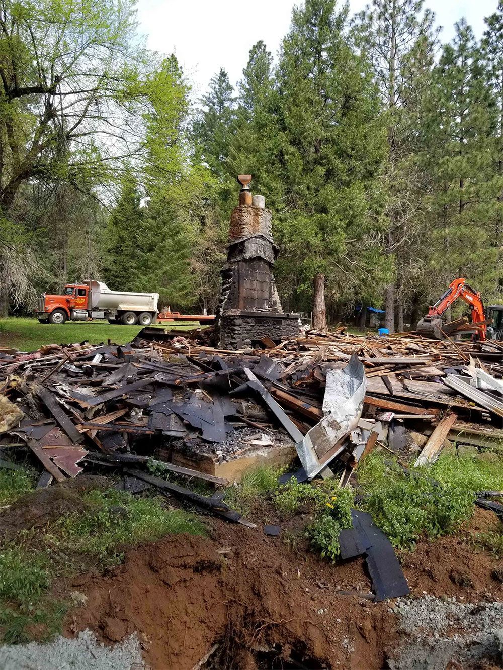 Debris of a destroyed building with a standing chimney. A dump truck and excavator are present in the background.