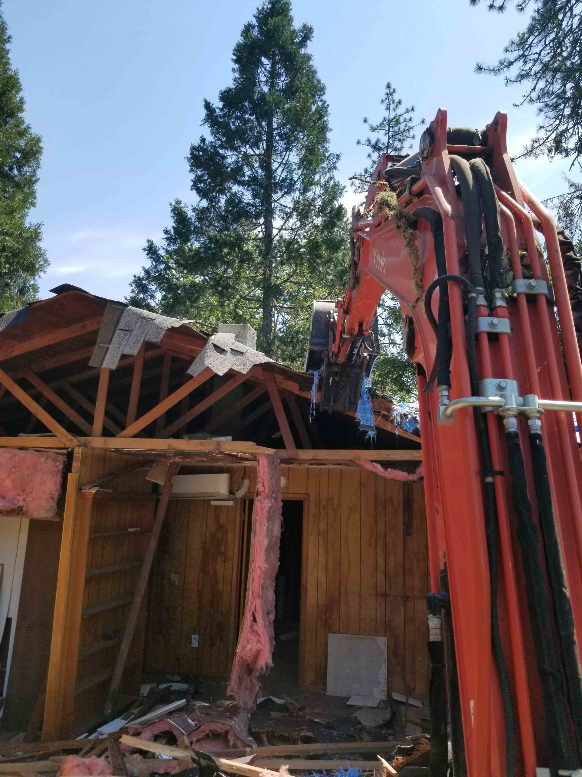 Excavator demolishing a building's roof; debris and insulation visible. Sunny day, trees in the background.