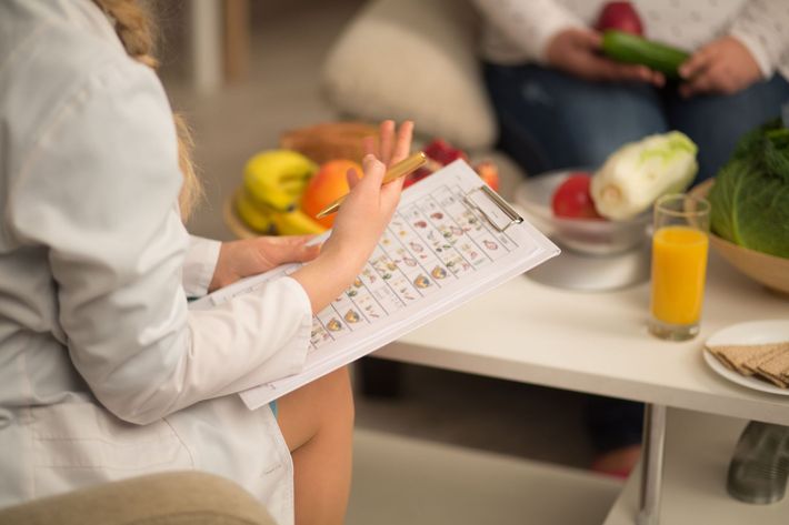 A woman is sitting at a table with fruits and vegetables and a clipboard.