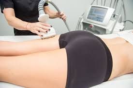 Woman receiving body contouring treatment on her thigh, with a machine and technician in a clinic.