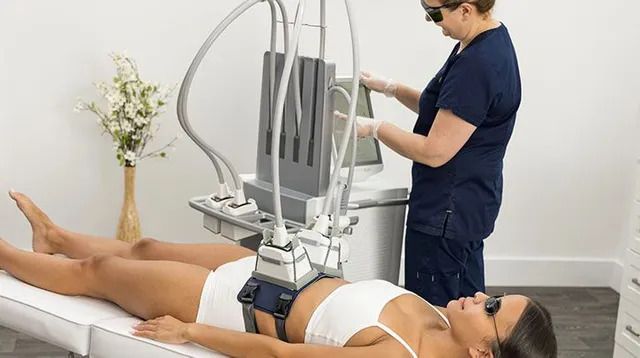 Woman receiving body sculpting treatment, lying on table, with technician operating machine. White room.
