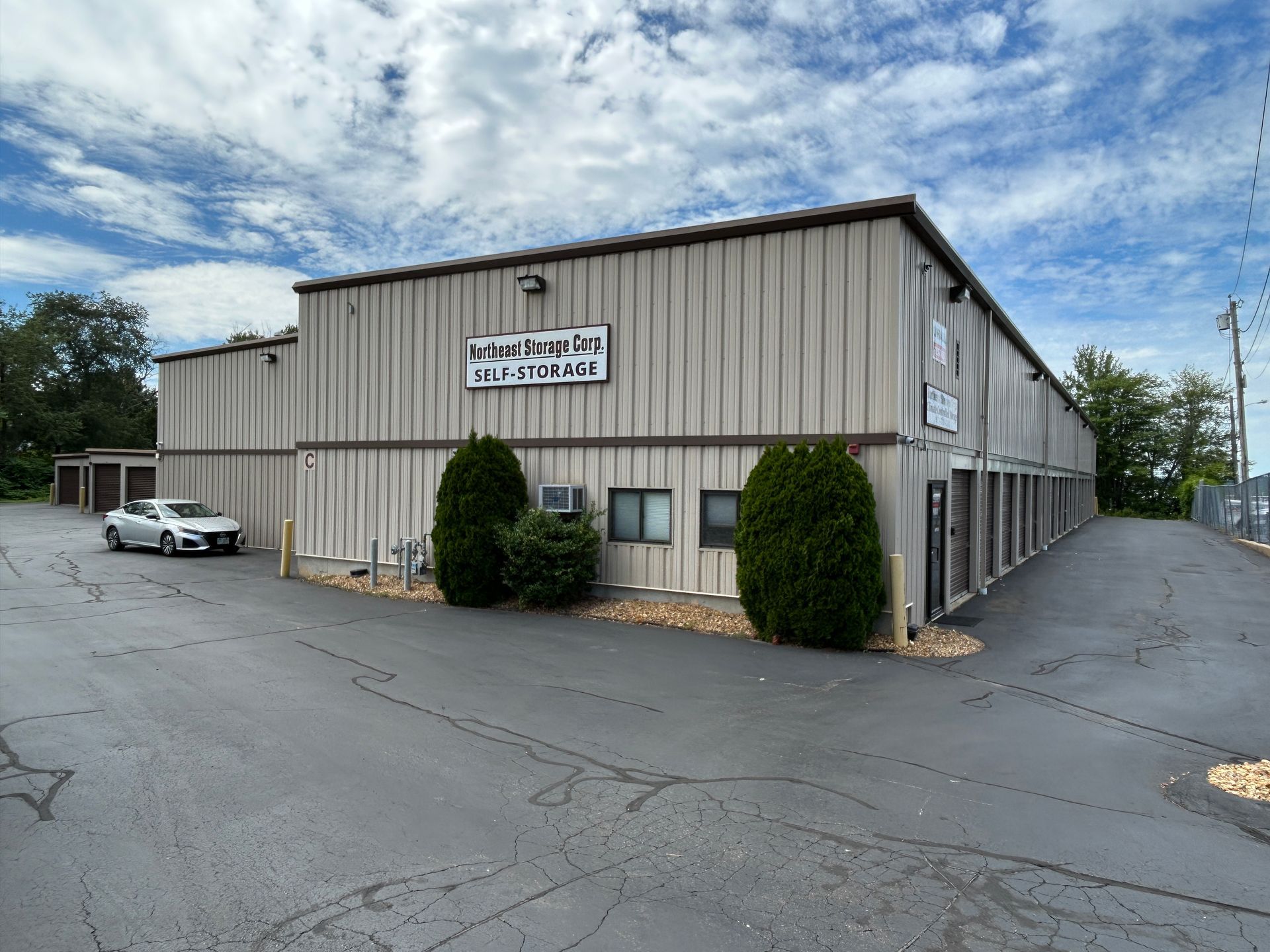 Gray industrial warehouse with a sign, parked car, and cloudy sky in a paved lot