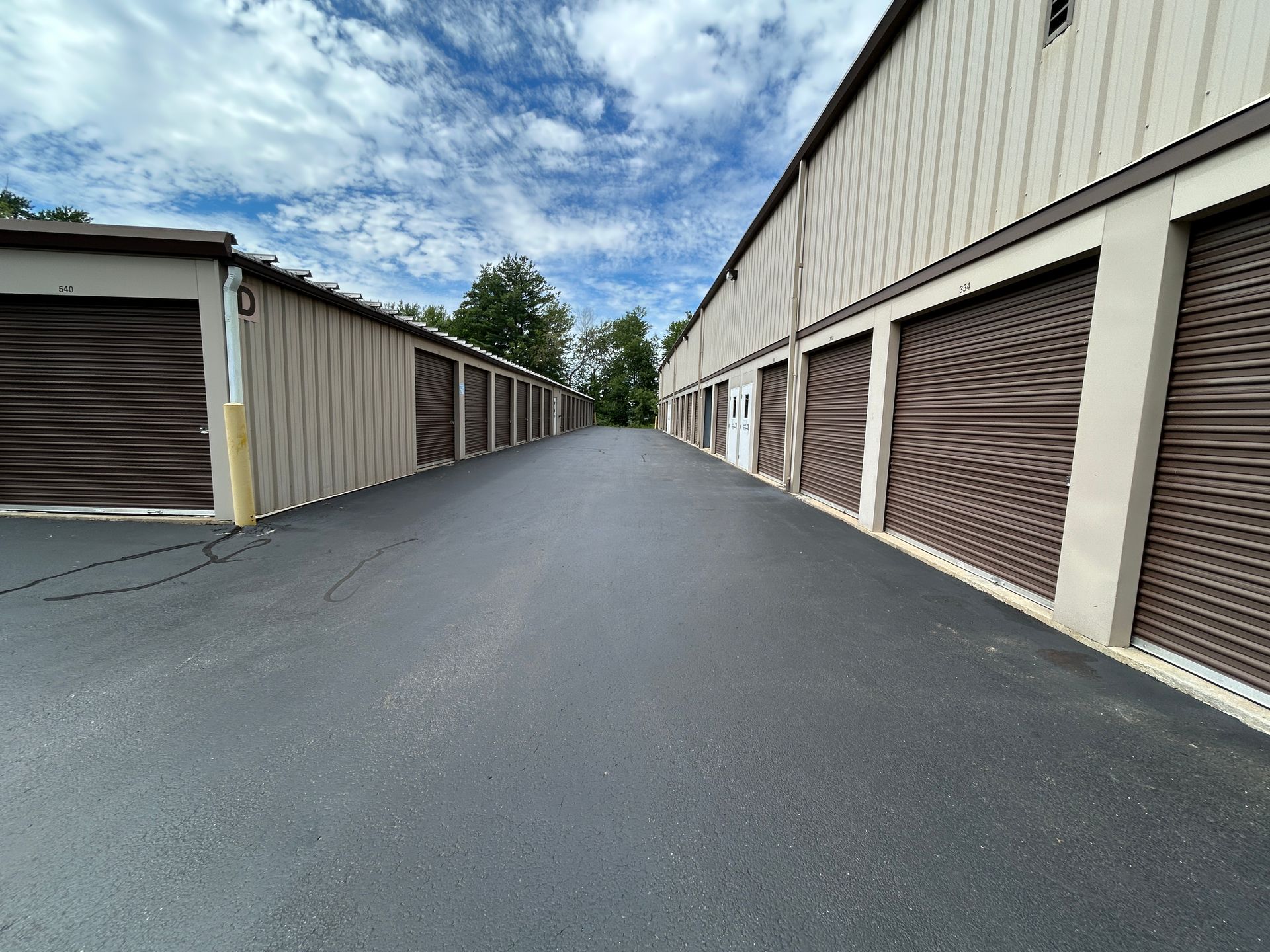 Storage unit row with closed brown doors along a paved lane under a partly cloudy sky