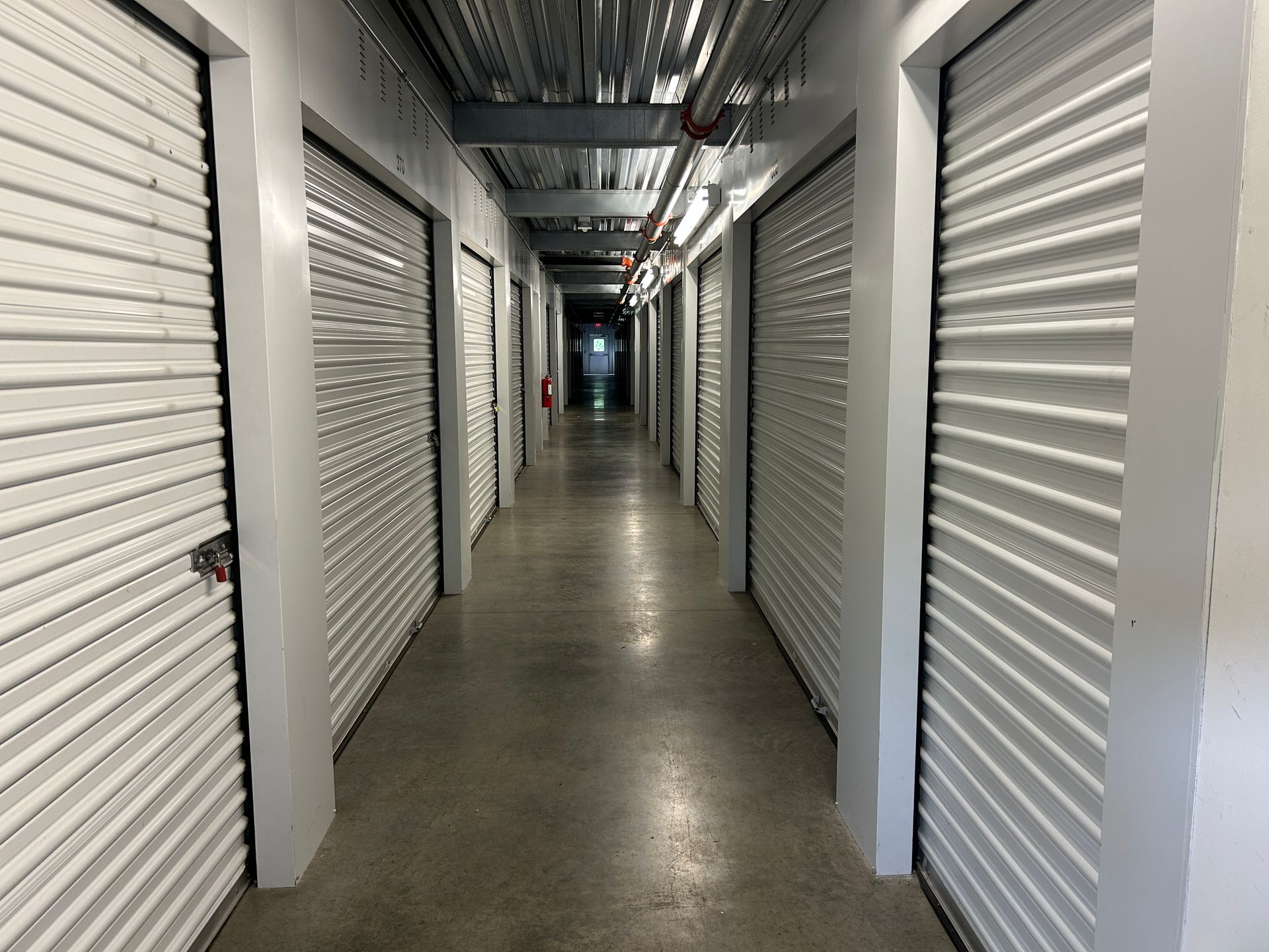 Indoor storage unit hallway with white roll-up doors and overhead pipes, receding into the distance.