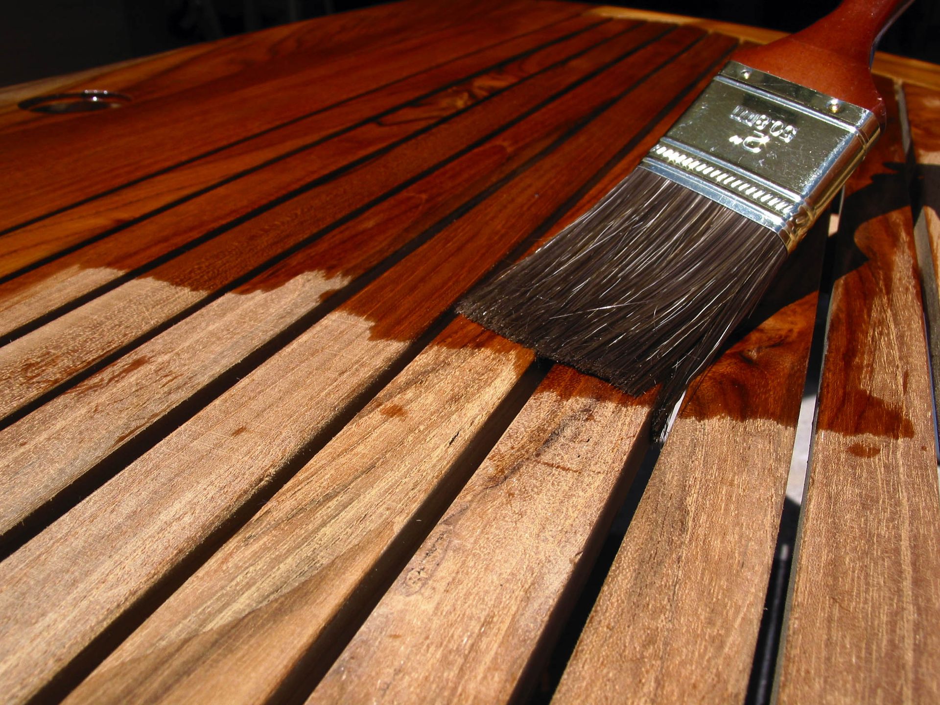 A paintbrush applying brown stain to wooden slats; half stained, half unstained.