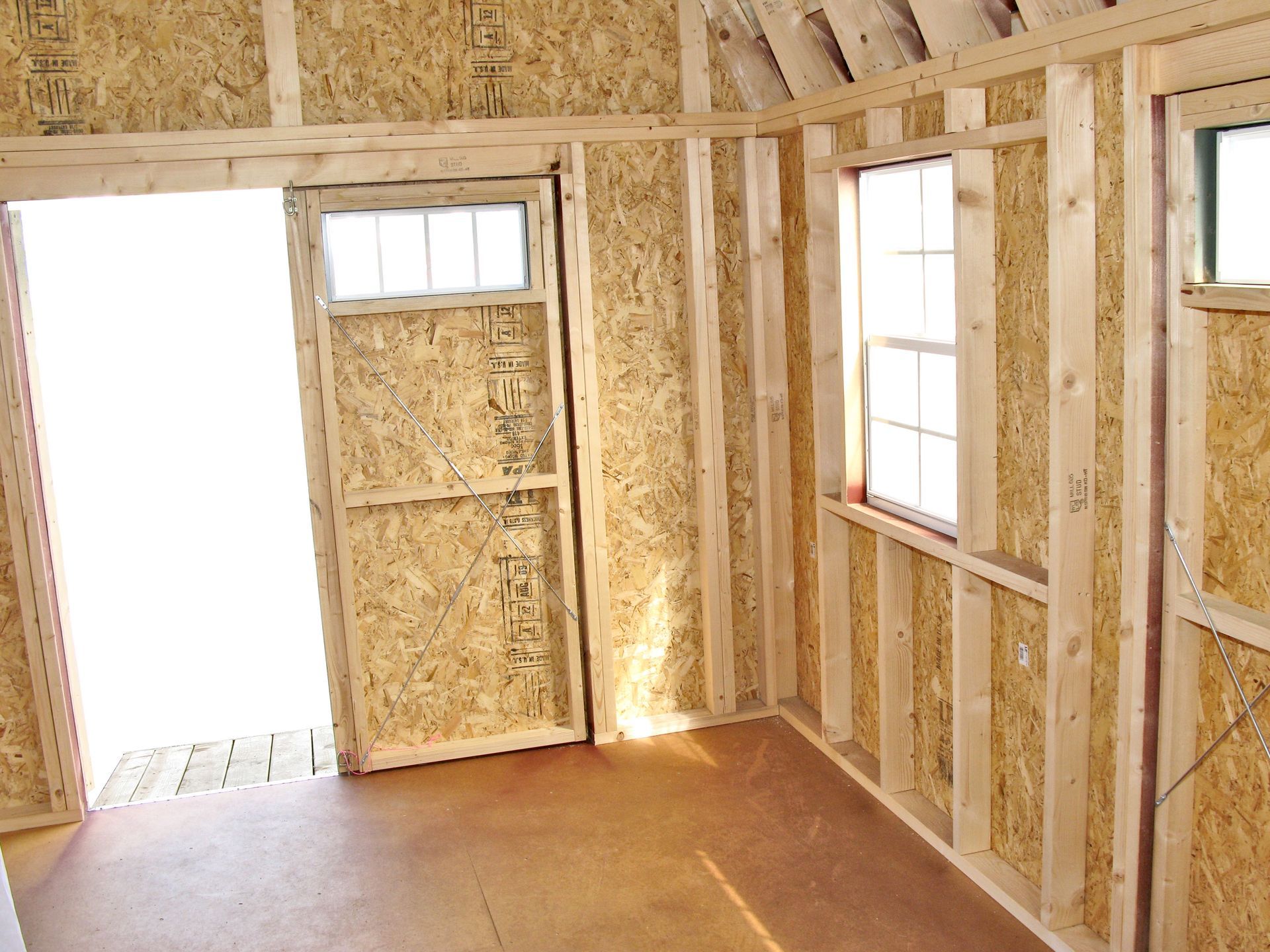 Interior of a wood-framed shed with a door, windows, and light brown floors and walls.