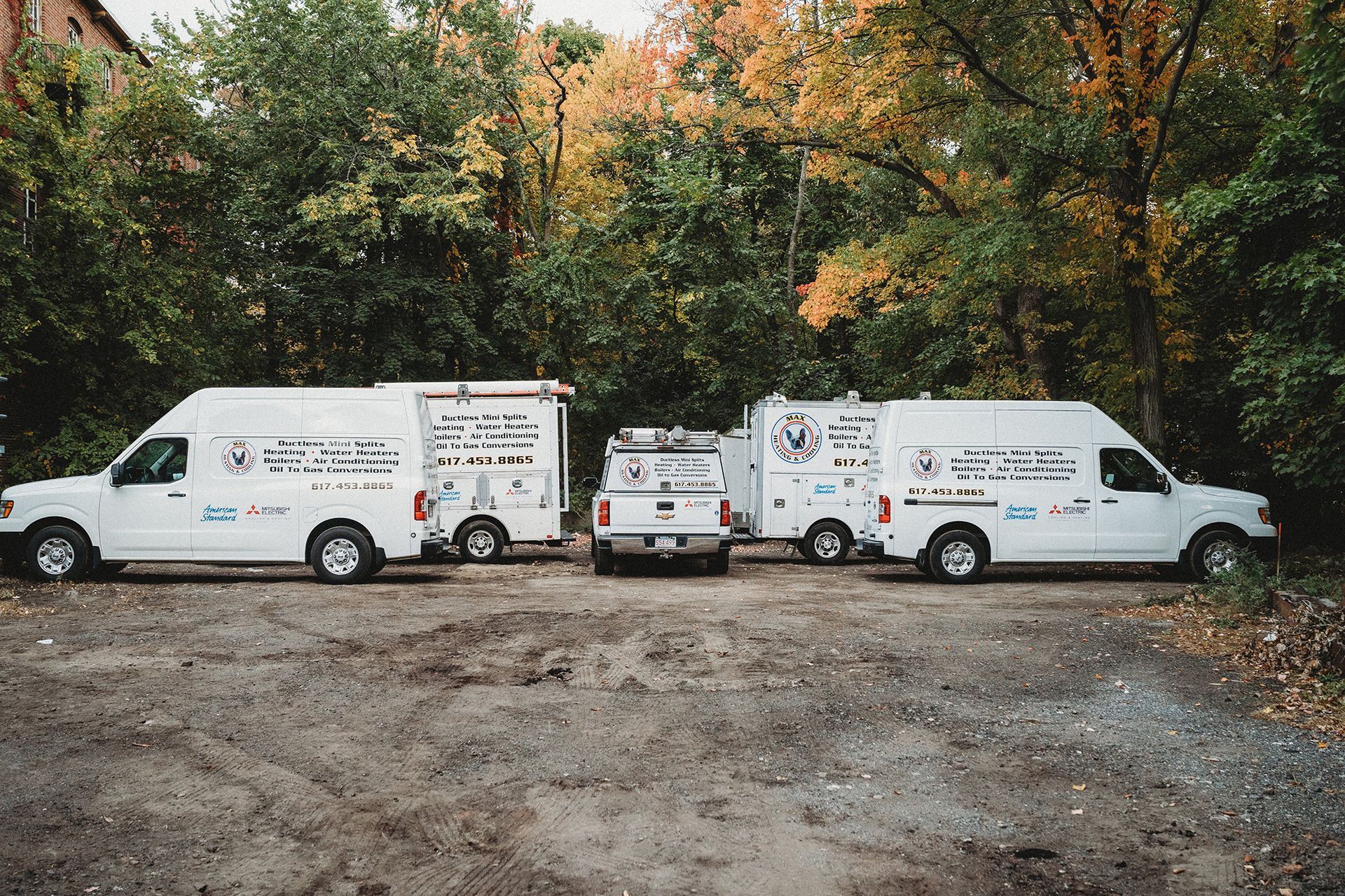 Three white service vans parked on a gravel lot with autumn trees in the background