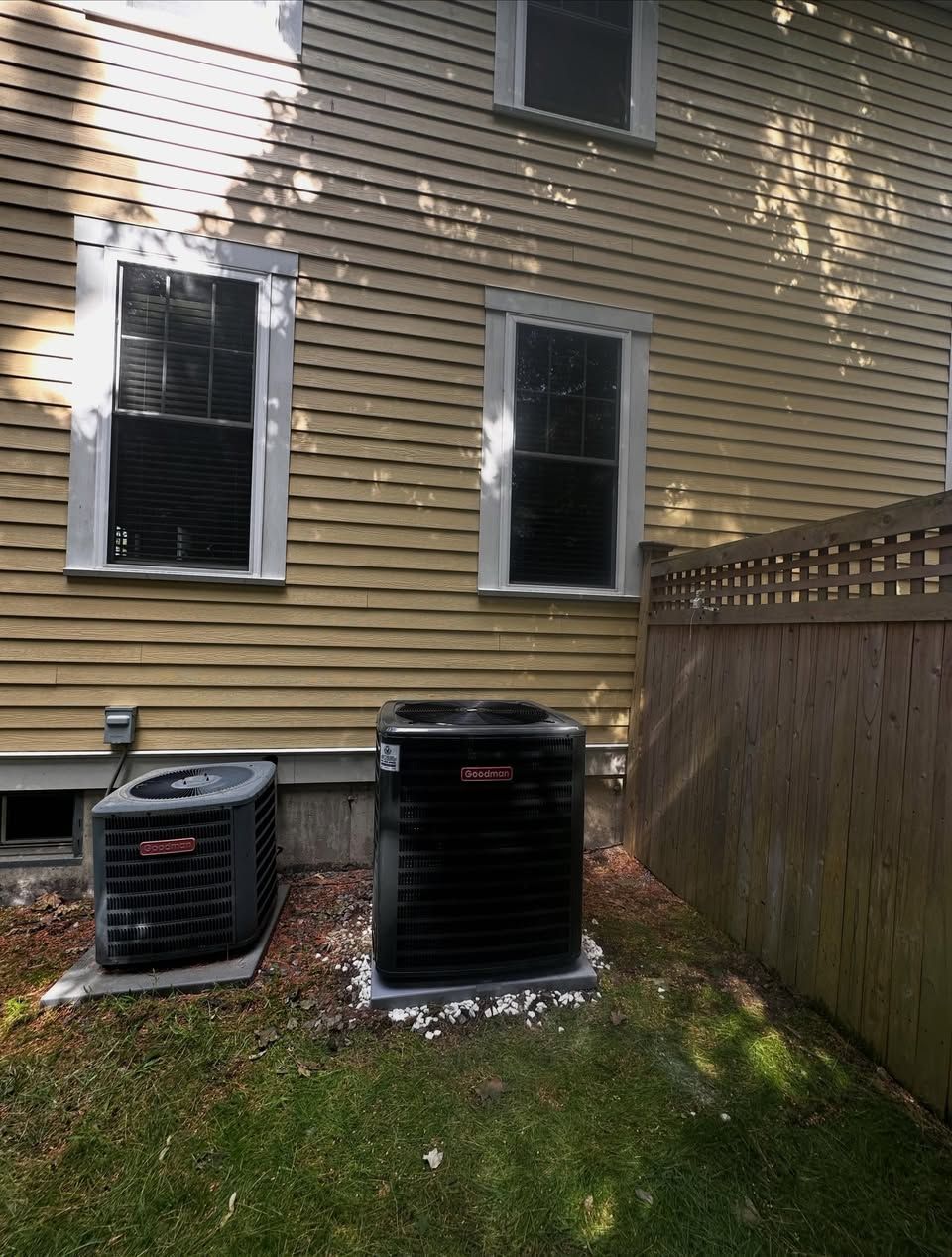Backyard beside a beige house with two windows, two air conditioners, grass, and a wooden fence.