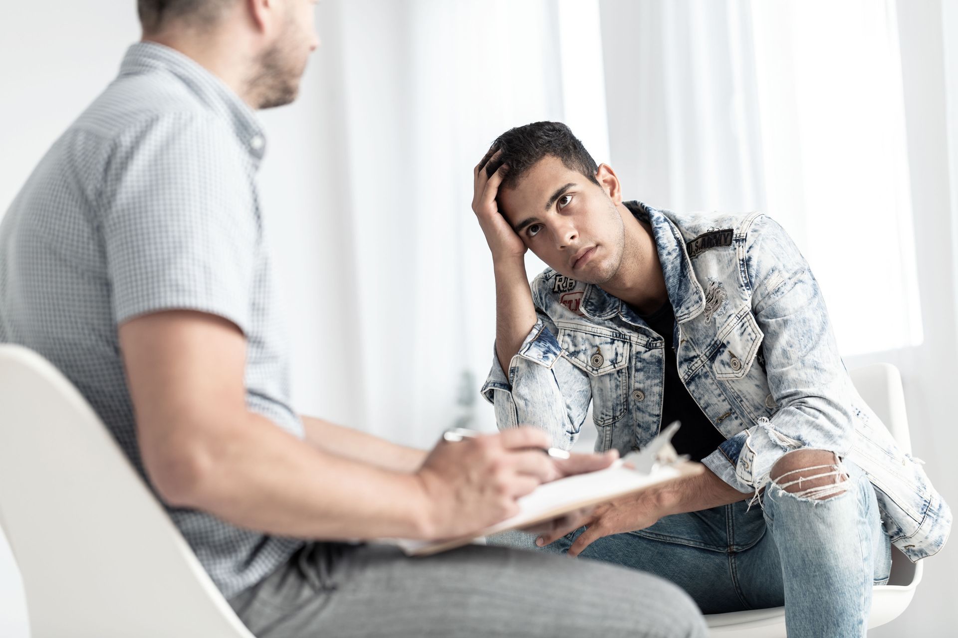 A smiling young person talks to a professional in a beige jacket holding a clipboard while sitting on a couch.
