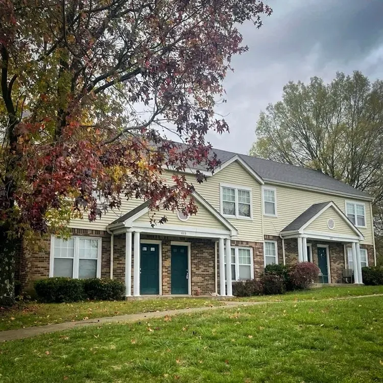 Apartment building with beige siding and green doors, framed by trees and a grassy lawn