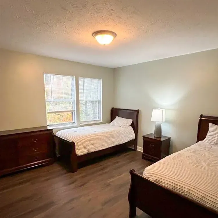 Spare bedroom with two dark wood twin beds, nightstand lamp, window, and dresser against beige walls.