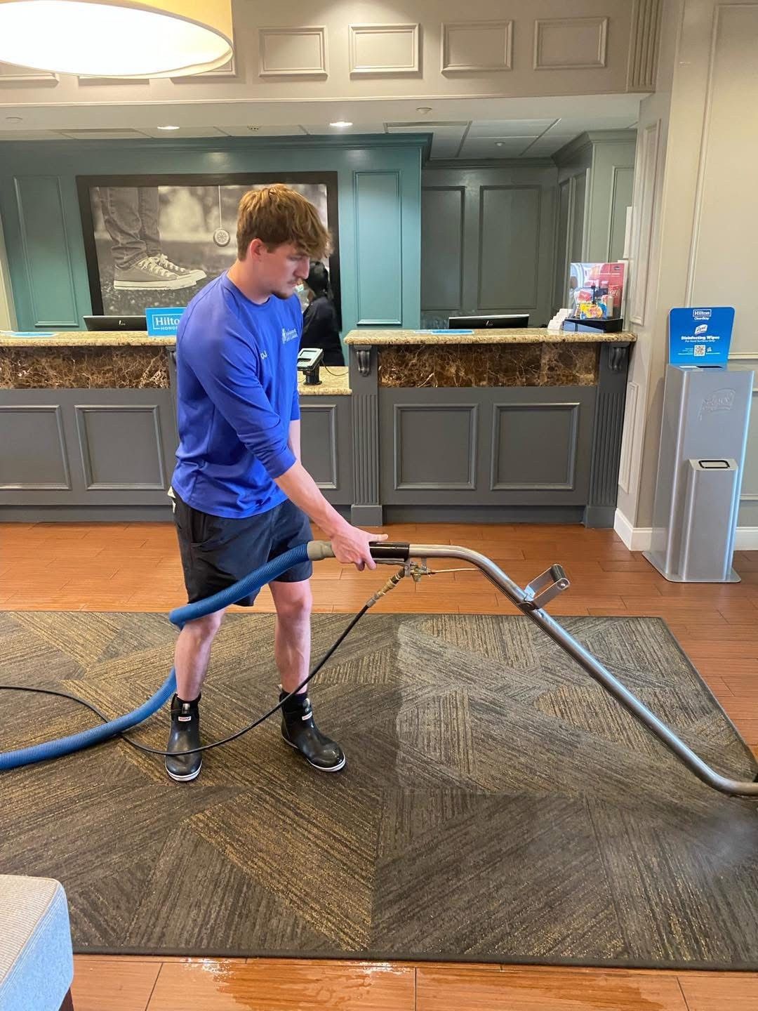 Person in blue shirt using a floor-cleaning machine on a carpeted lobby floor.