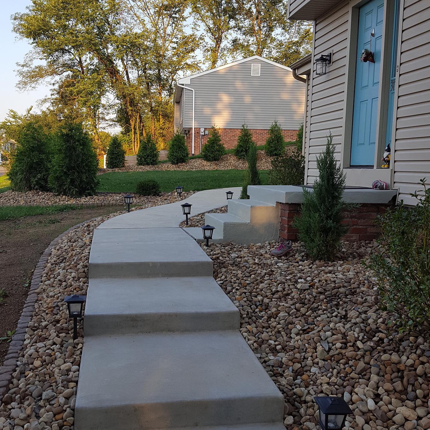 Concrete walkway with steps leading to a blue front door beside a house, bordered by gravel and landscaping lights.