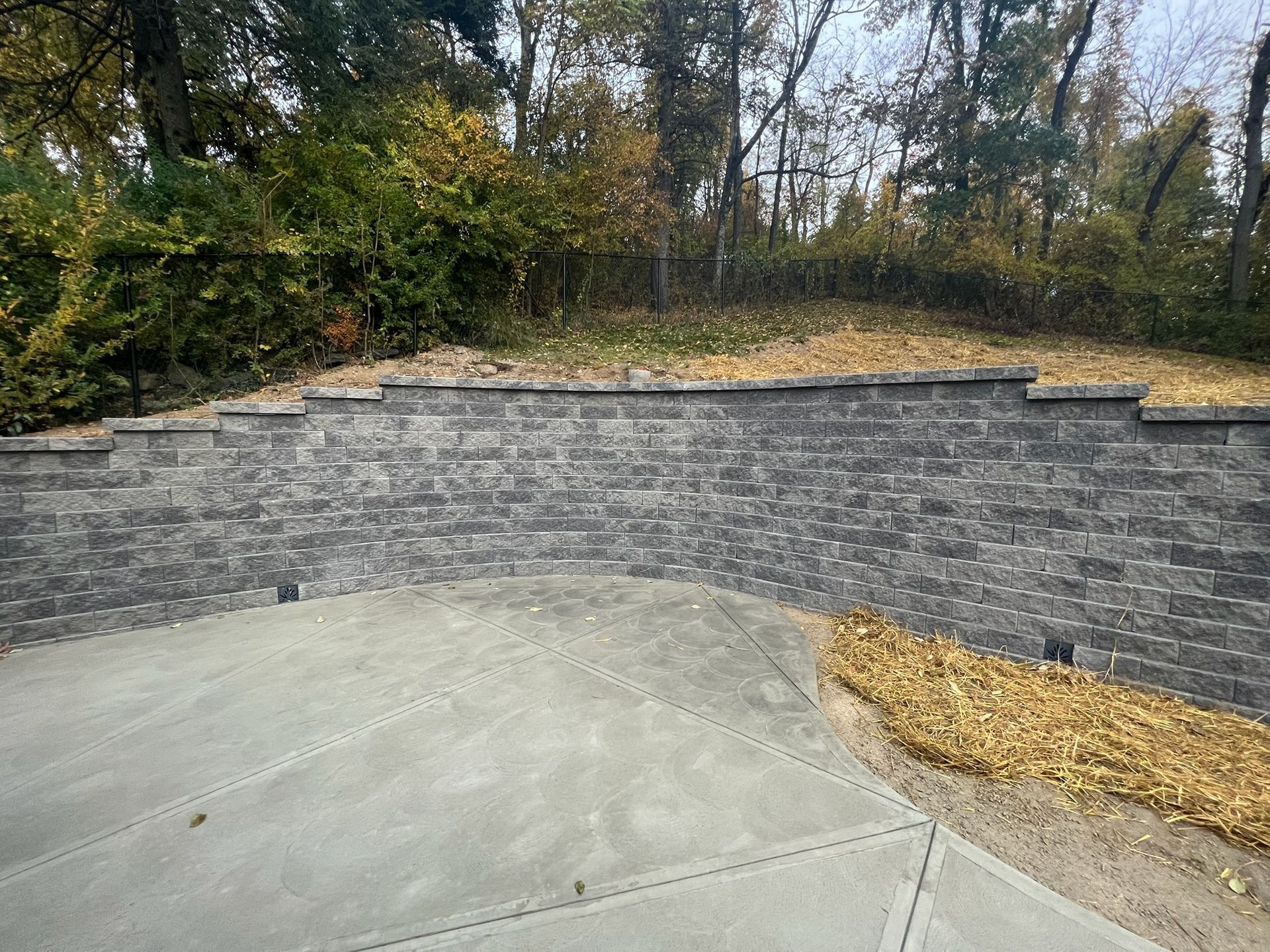 Gray stone retaining wall curving around a concrete patio beside a wooded hillside with fallen leaves