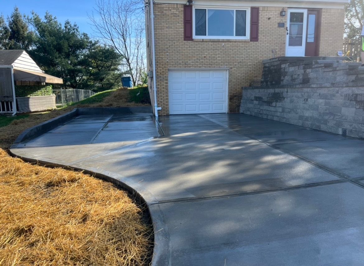 Concrete driveway leading to a garage beside a beige house, with a stone retaining wall and lawn edge.