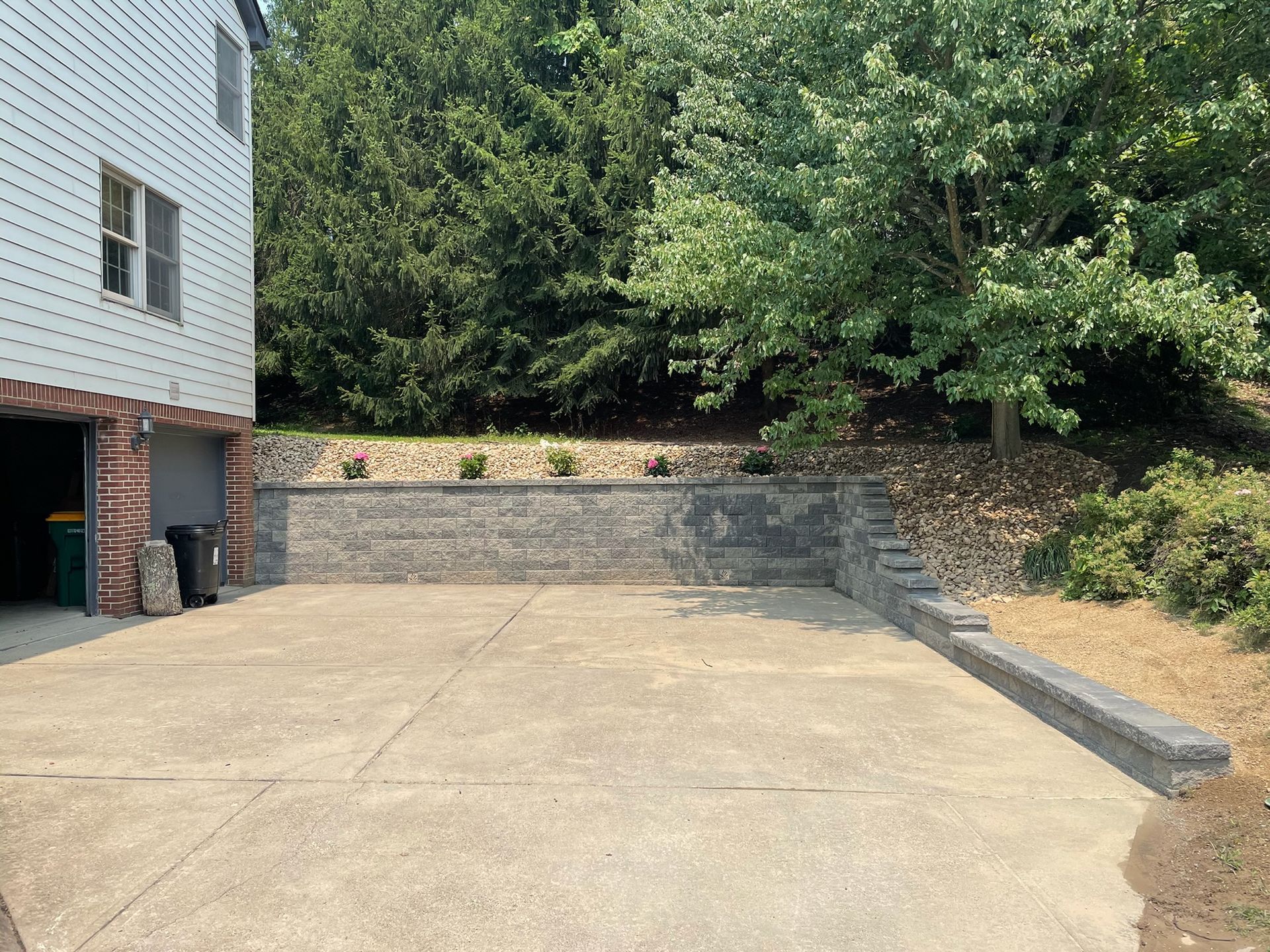 Concrete driveway beside a house, sloping up to a stone retaining wall and trees.