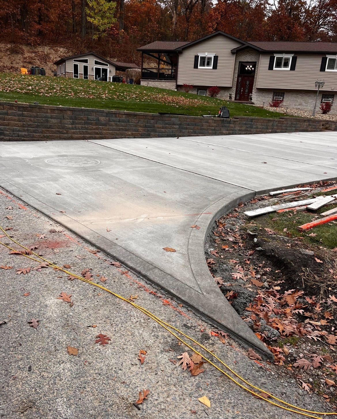Fresh concrete driveway beside a stone retaining wall and house in a leaf-covered residential yard.