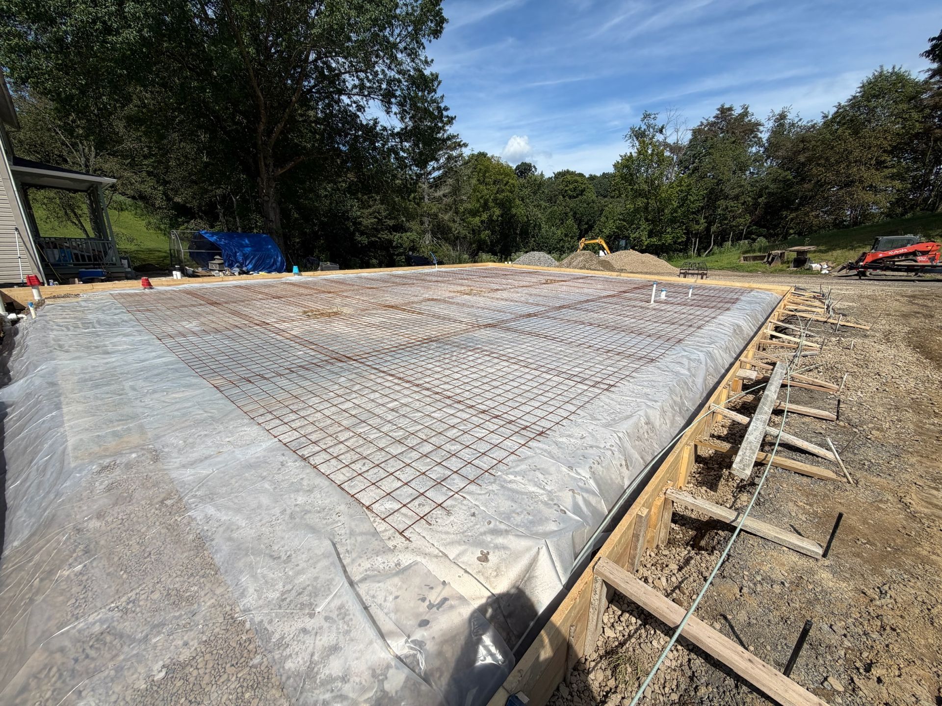 Concrete slab foundation under construction with rebar grid and plastic sheeting in a wooded area