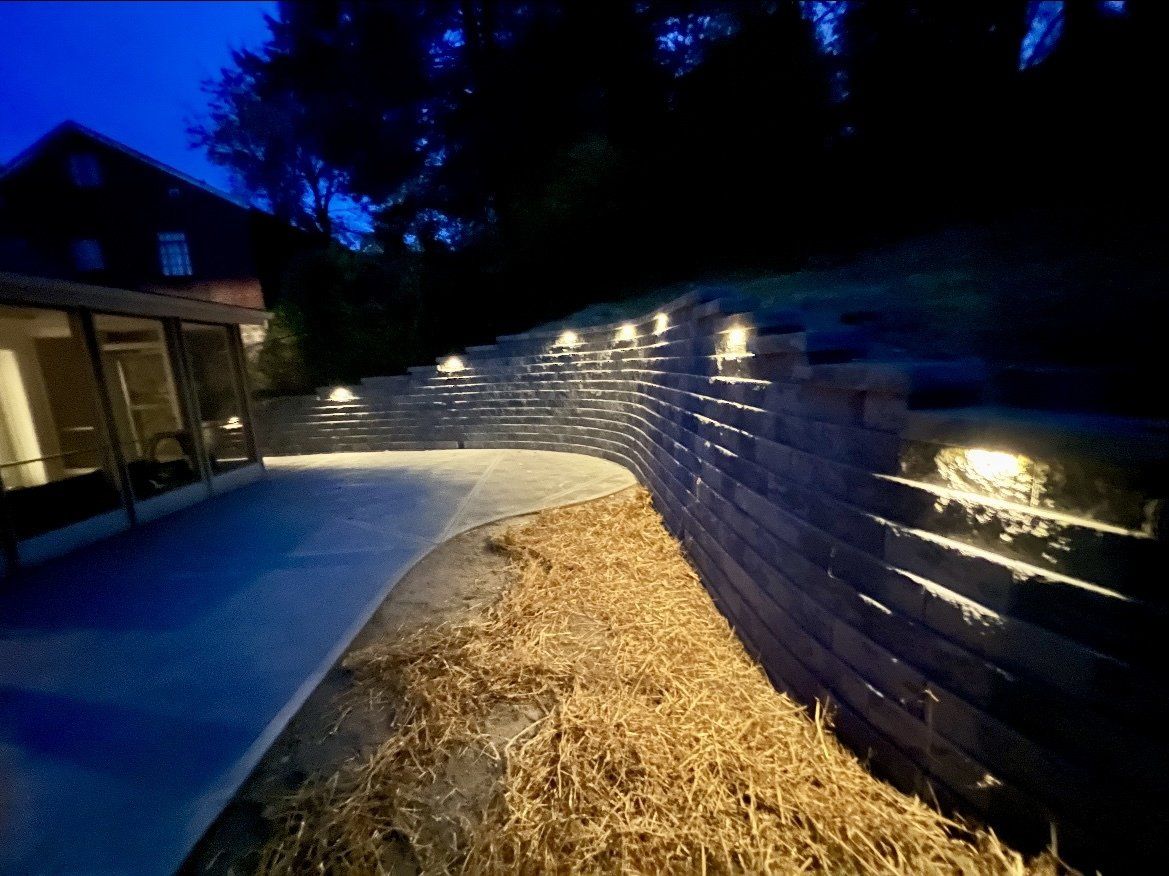 Curved backyard stone wall lit at dusk beside a house, with straw-lined path and blue evening sky