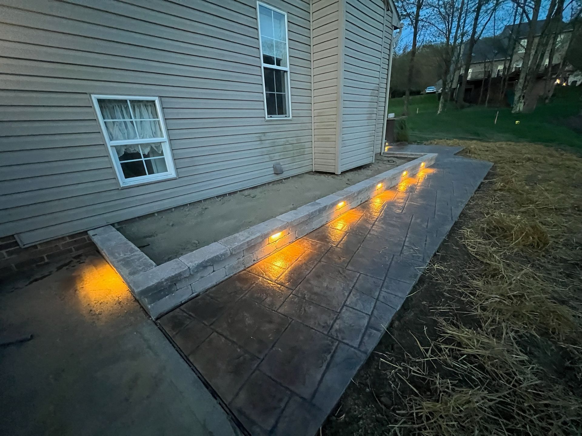 Flames burning along the edge of a concrete walkway beside a beige house at dusk