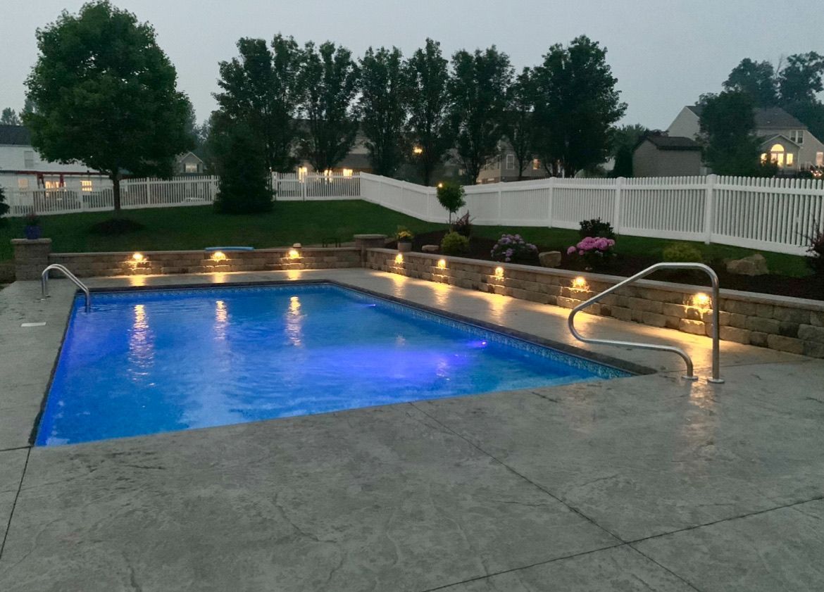 Backyard pool at dusk with blue-lit water, stone patio, and string lights along the fence
