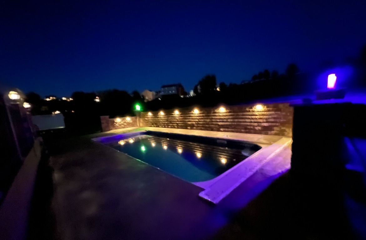 Lit backyard pool at night with warm string lights and a glowing blue water surface