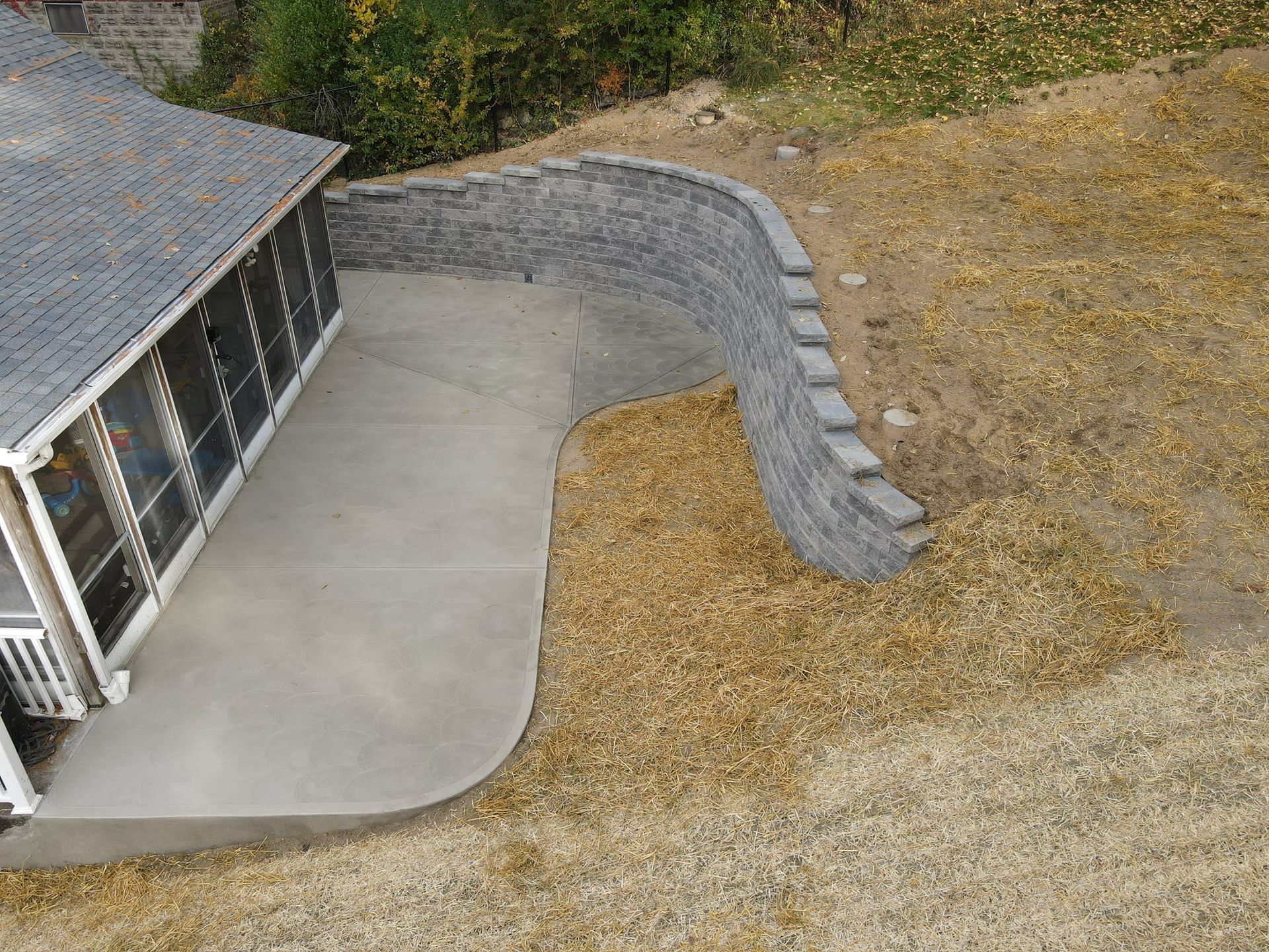 Concrete patio beside a house, bordered by a curved gray retaining wall and dry grass.