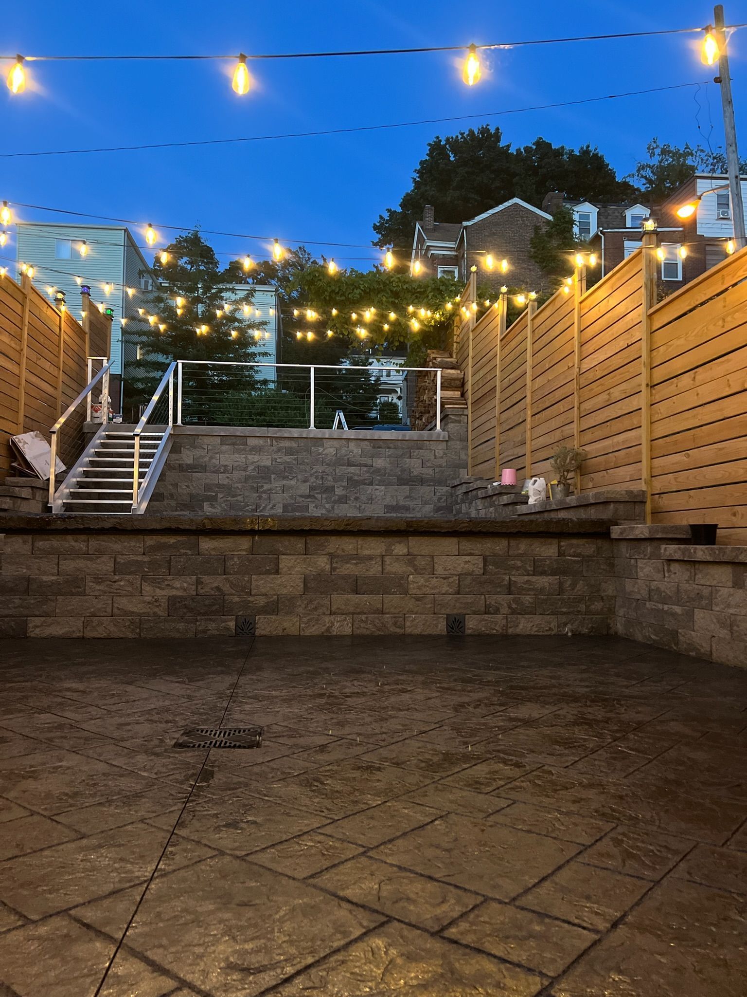 Twilight patio with string lights, stone steps, and wooden fencing leading to a higher terrace