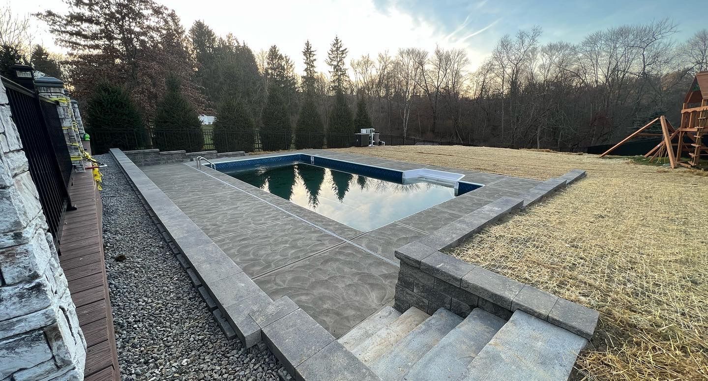Backyard swimming pool surrounded by gravel and concrete steps, with trees in the background.
