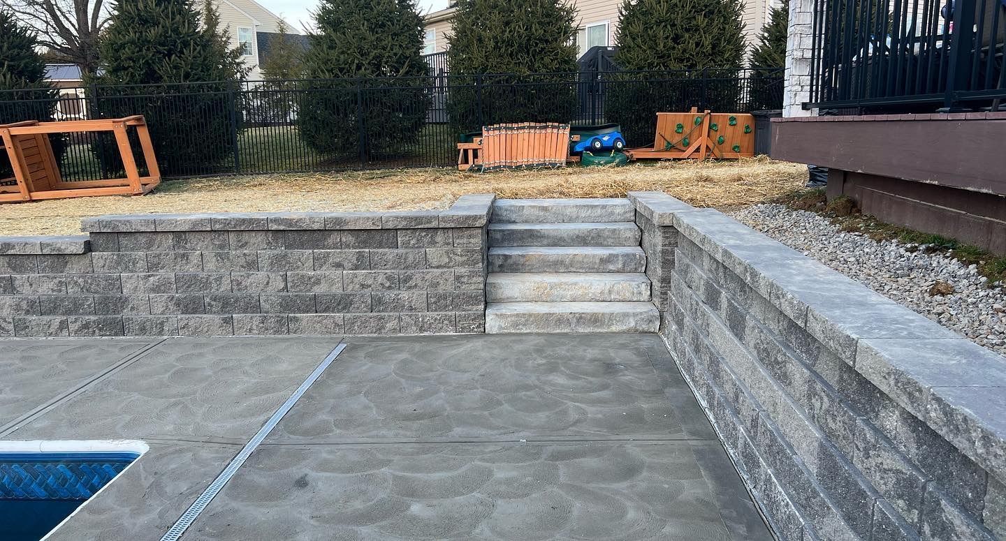 Empty concrete pool deck with stone retaining walls and steps in a backyard.