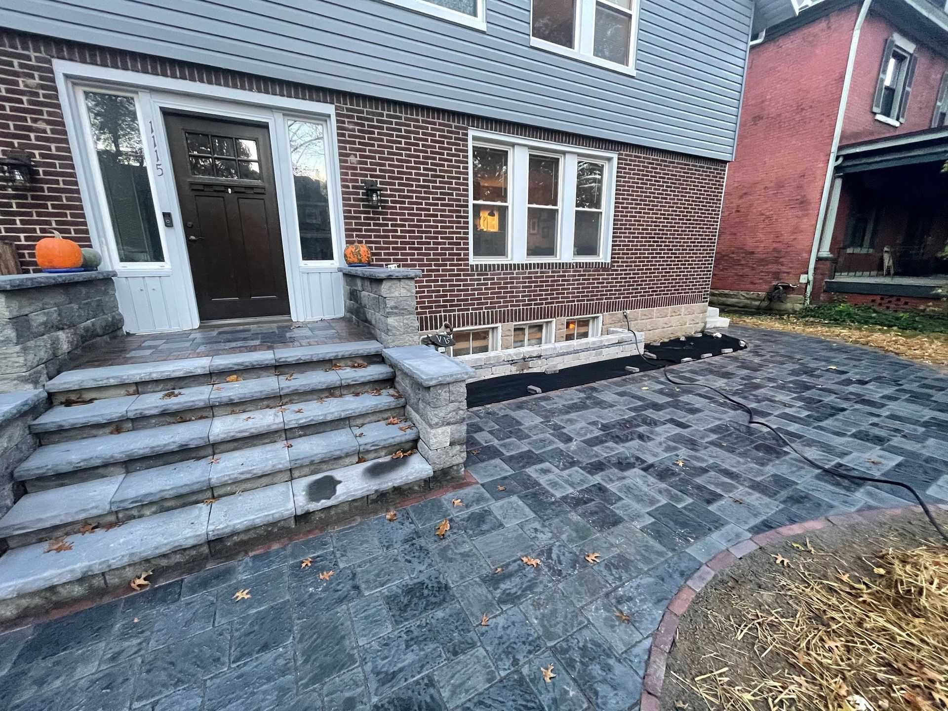 Front entry with stone steps and black paver patio beside a brick house, pumpkins by the door.