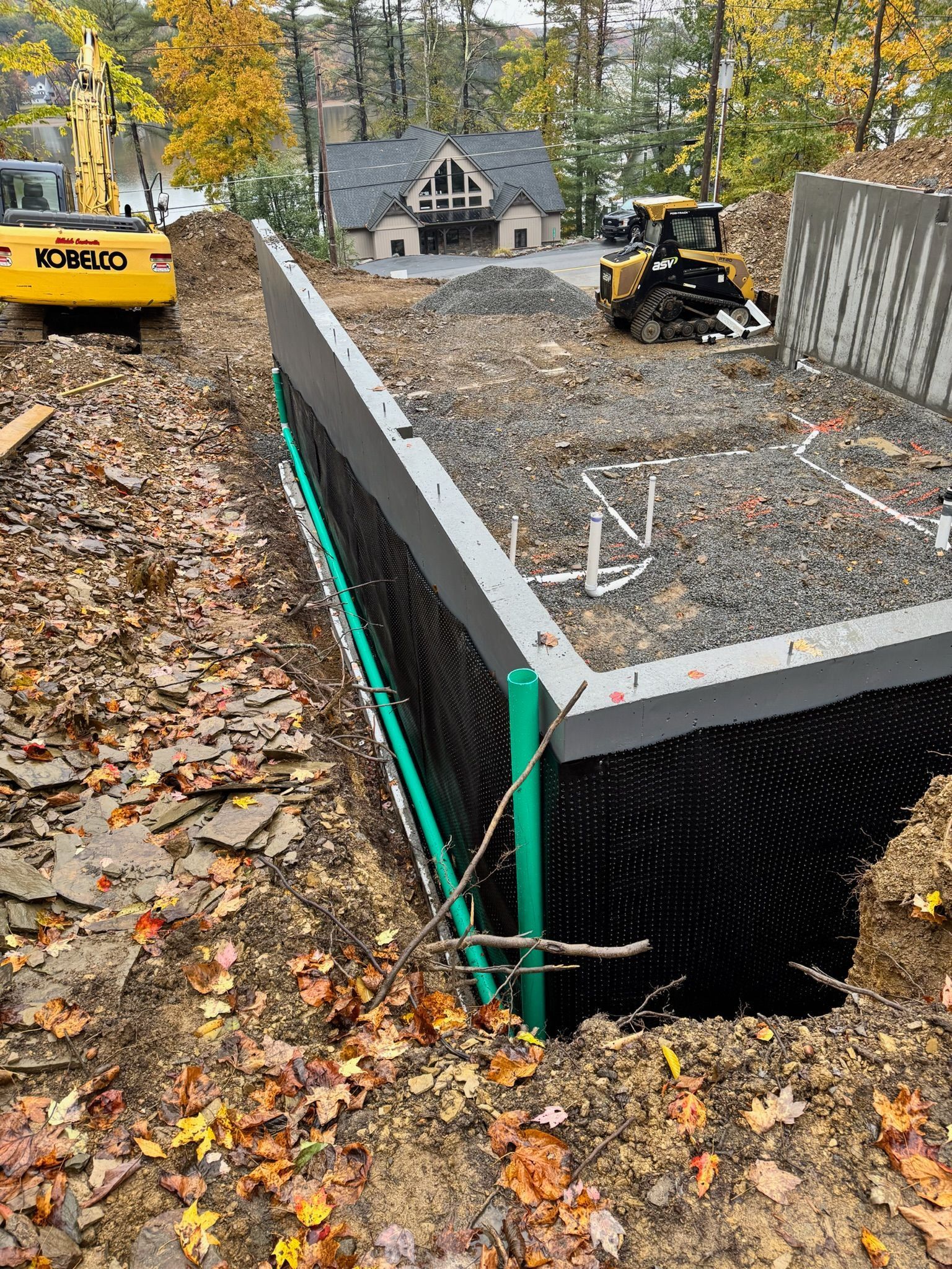 Construction site with excavator and retaining wall over a dug-out foundation pit in a wooded residential area