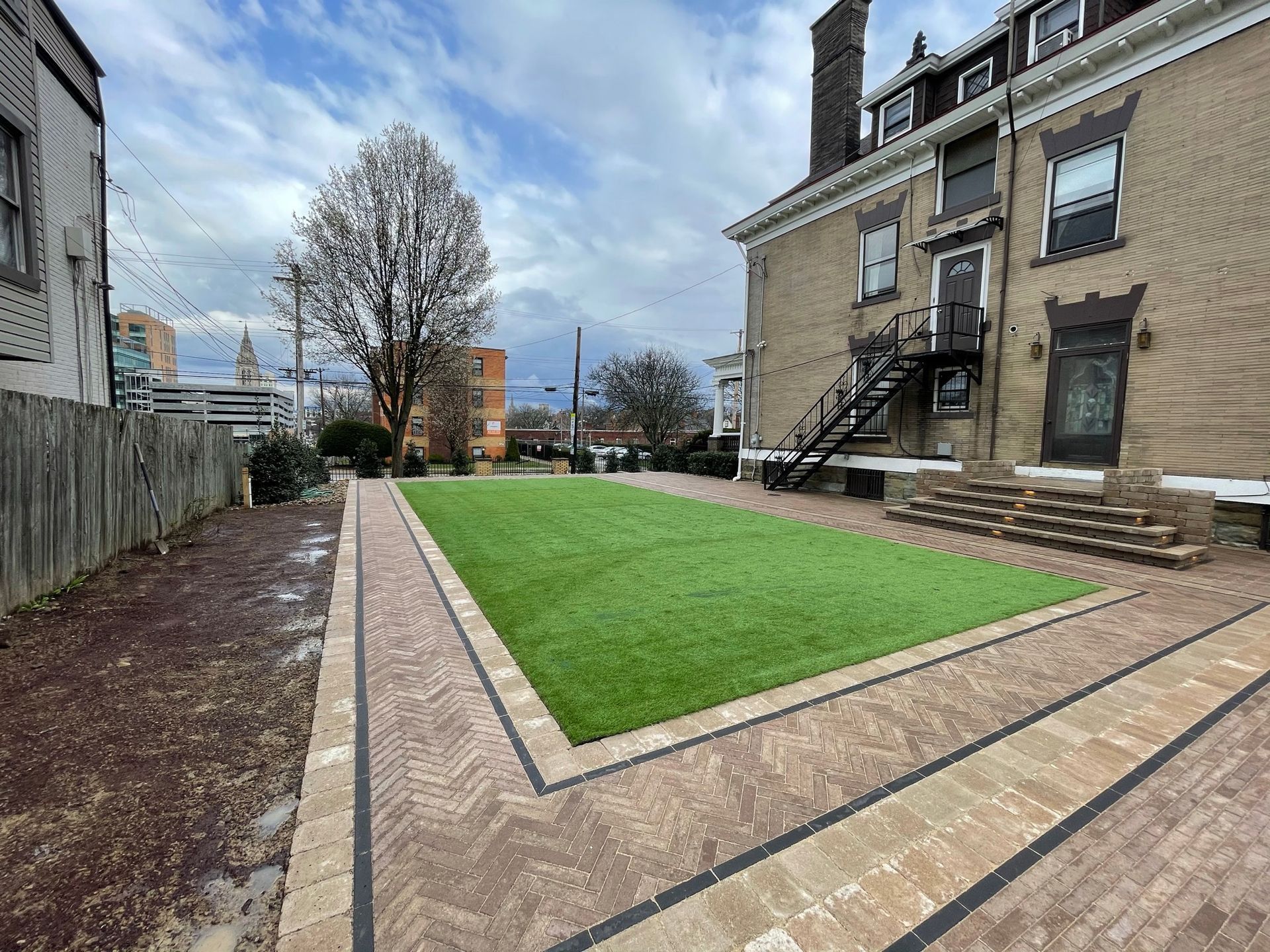 Landscaped courtyard with green lawn, brick paths, and a stone building beside a fenced garden area.