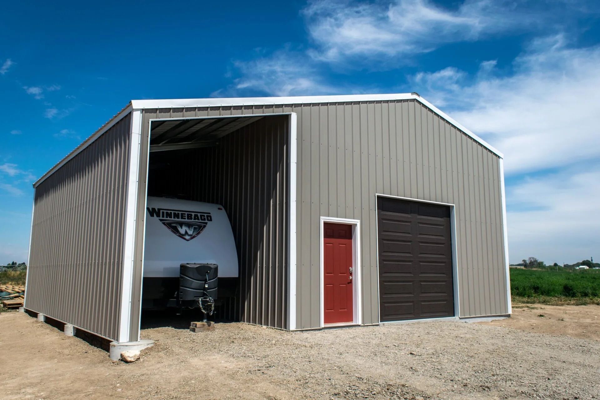 Large gray metal shed with red and black doors on a gravel lot under a blue sky