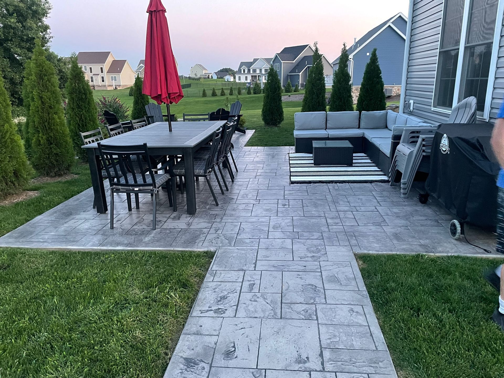 Patio with dining table, red umbrella, sectional seating, grill, and stone pavers in a fenced backyard.
