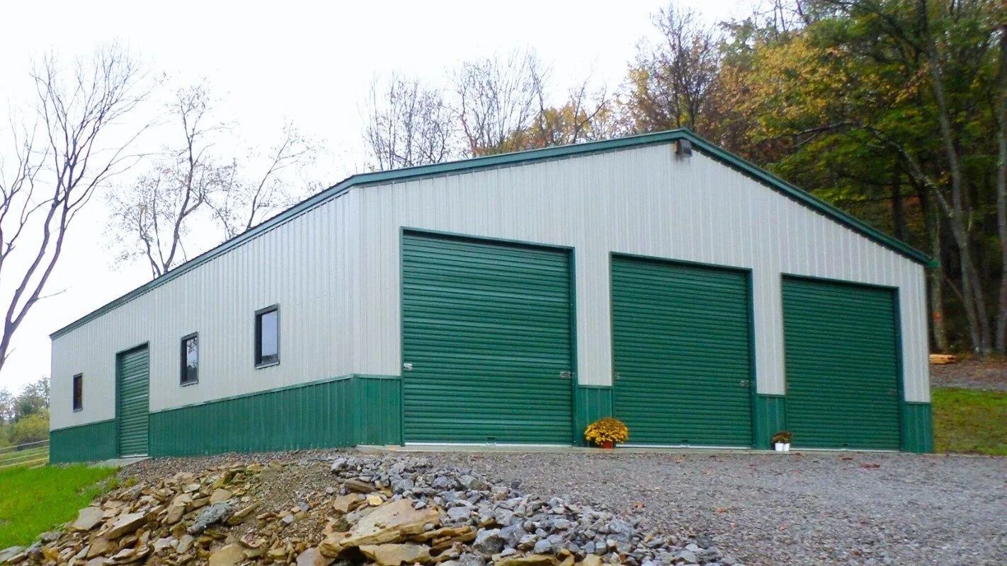 Green-and-white metal storage building with large garage doors on a gravel lot, surrounded by trees