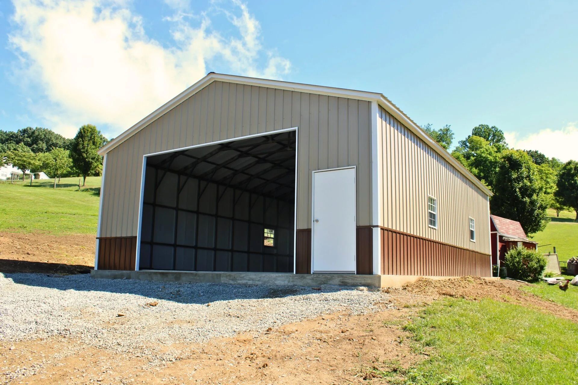 Large tan metal barn with open front door on a grassy hillside under a blue sky