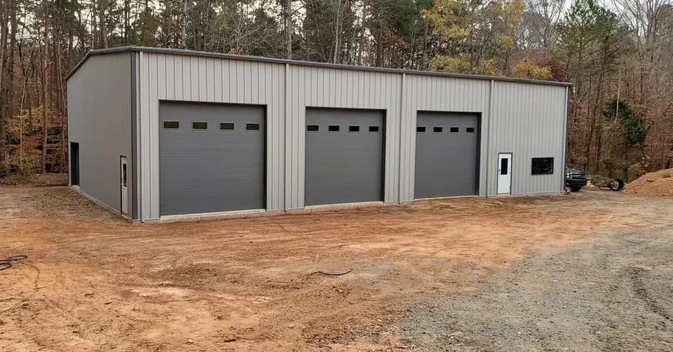 Gray industrial garage building with four roll-up doors on a dirt lot surrounded by trees