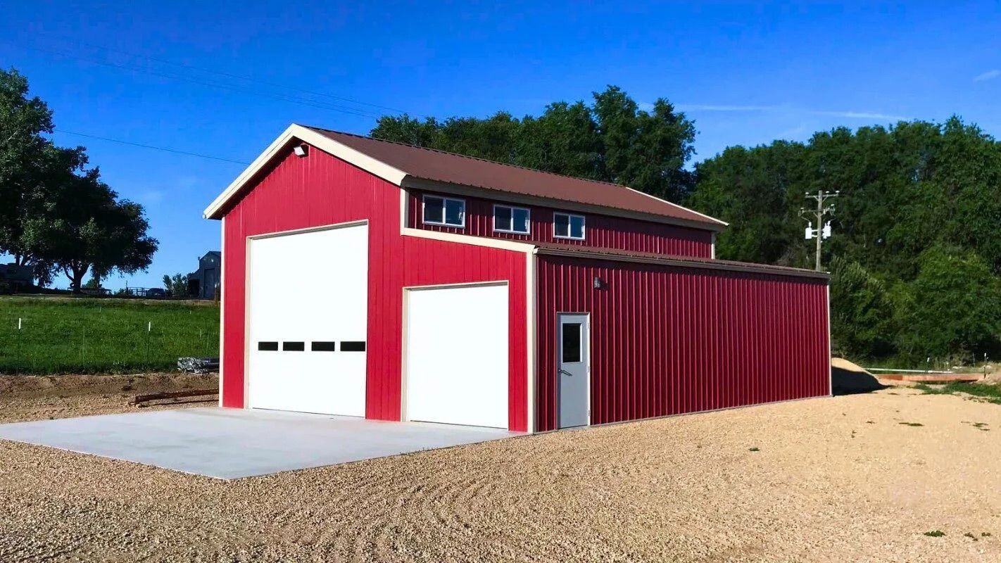 Red barn-style metal building with white garage door on a gravel lot, surrounded by trees.