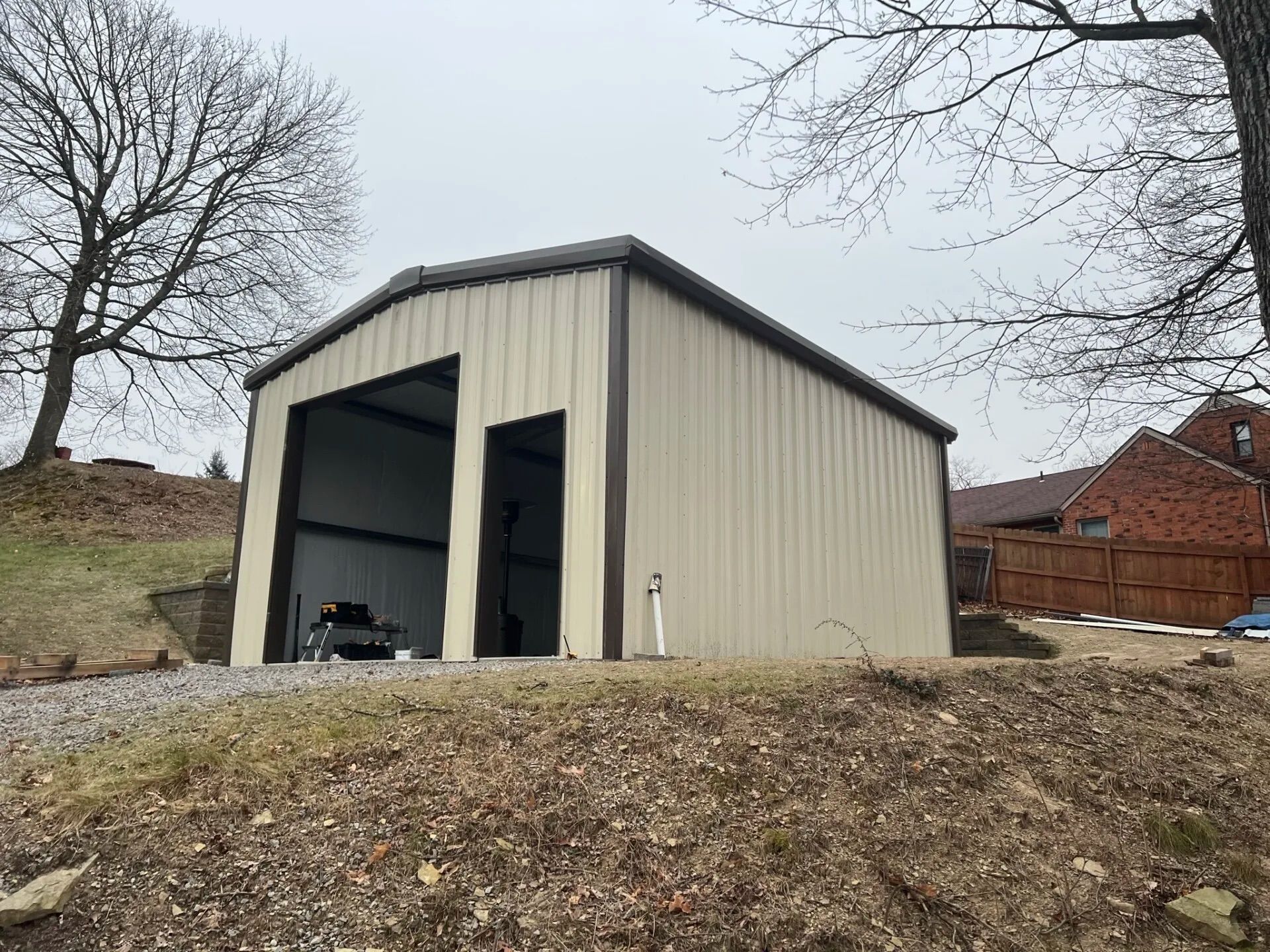 Large beige metal storage building on a dirt hill with open bay doors, surrounded by bare trees.
