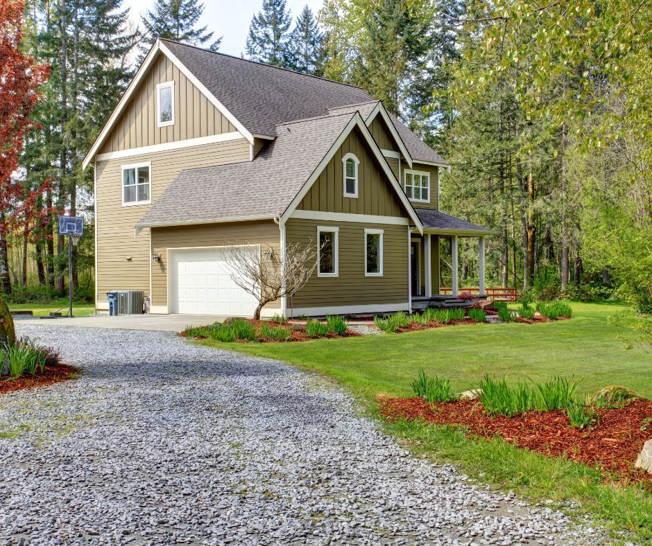 Two-story tan house with garage, gravel driveway, and green lawn beside trees