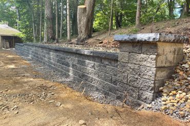 Stone retaining wall along a wooded hillside with exposed soil and stacked rocks at the end