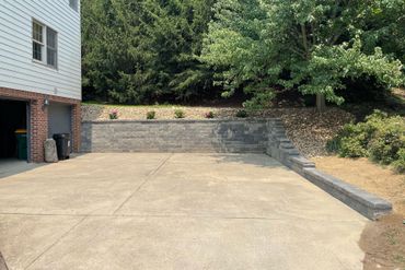 Concrete driveway beside a house, leading up to a stone retaining wall and trees