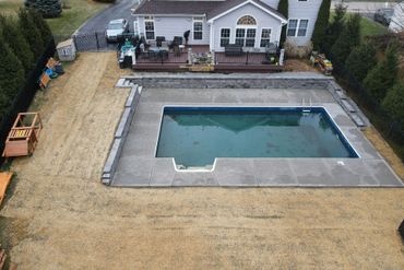 Backyard with a rectangular in-ground pool beside a house and patio, surrounded by dry grass and trees