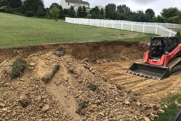 Red skid steer reshaping a dirt embankment beside a grassy field and white fence