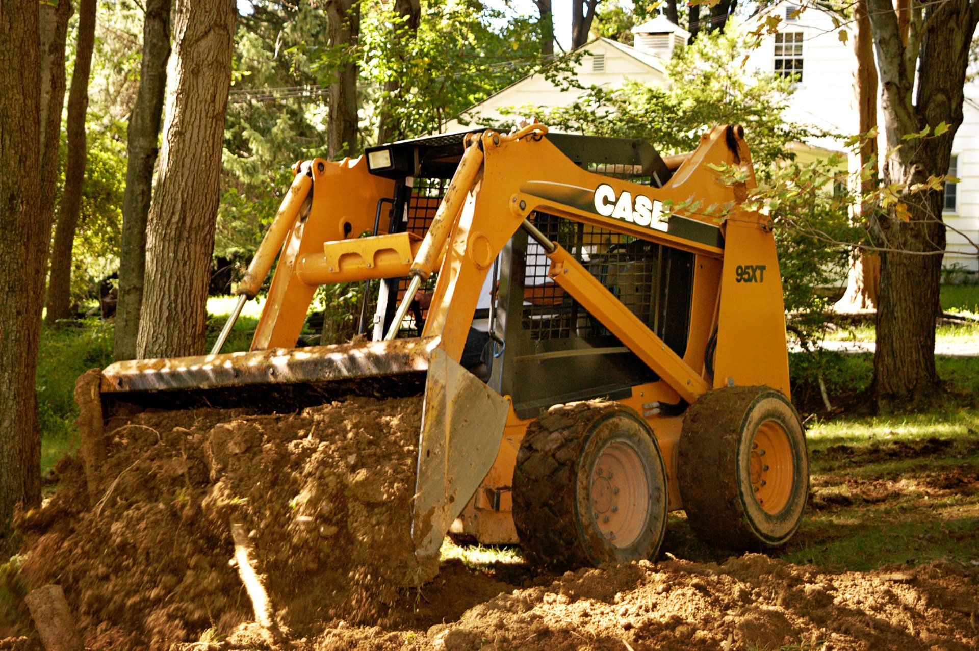 Yellow skid steer loader pushing dirt in a wooded area with trees and a house in the background