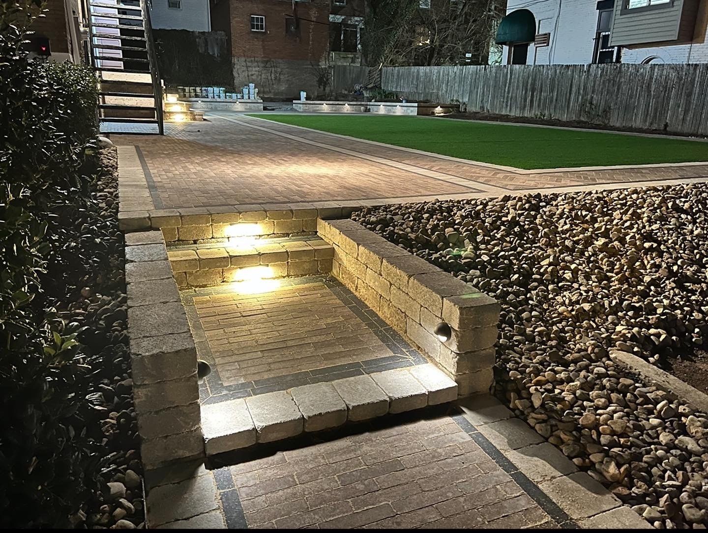 Lit stone steps leading to a patio, with ground lights and gravel landscaping at night.